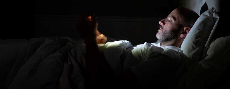 En person som ligger i en seng med en mobiltelefon i hånden som lyser opp ansiktet i mørket, søvn