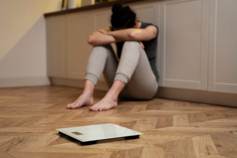 En trist kvinne sitter på gulvet og dekker hodet med armene sine, med en vekt foran. Illustrasjonsbilde for spiseforstyrrelse
